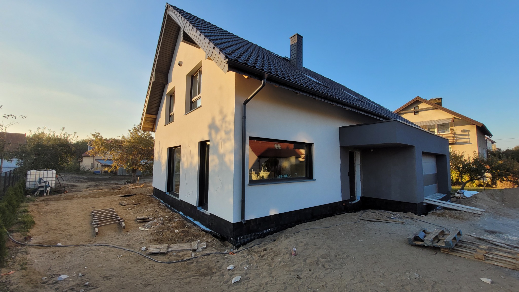 Nowoczesny dom murowany z szarą elewacją, czarną dachówką i oknami, widok z zewnątrz w trakcie wykańczania terenu wokół budynku.