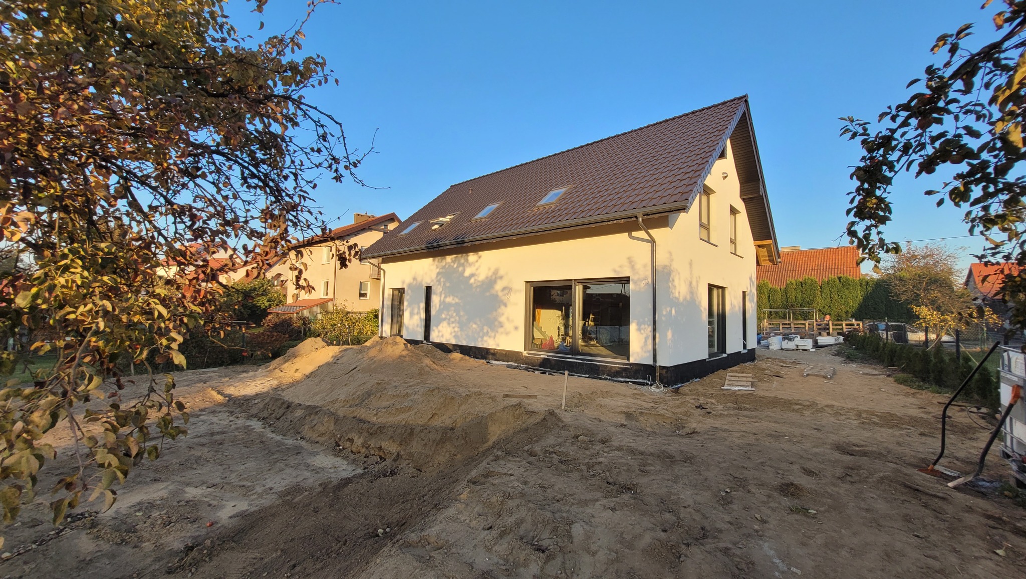 Nowo wybudowany, parterowy dom z białymi ścianami i brązowym dachem, widoczne okna dachowe, teren wokół domu w trakcie przygotowania, hałdy piasku i ziemi.