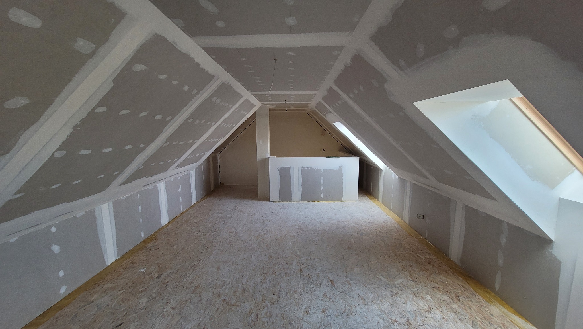 Poddasze w trakcie adaptacji: surowe ściany z płyt gipsowo-kartonowych z widocznymi łączeniami, podłoga z płyt OSB, okno dachowe wpuszczające naturalne światło.