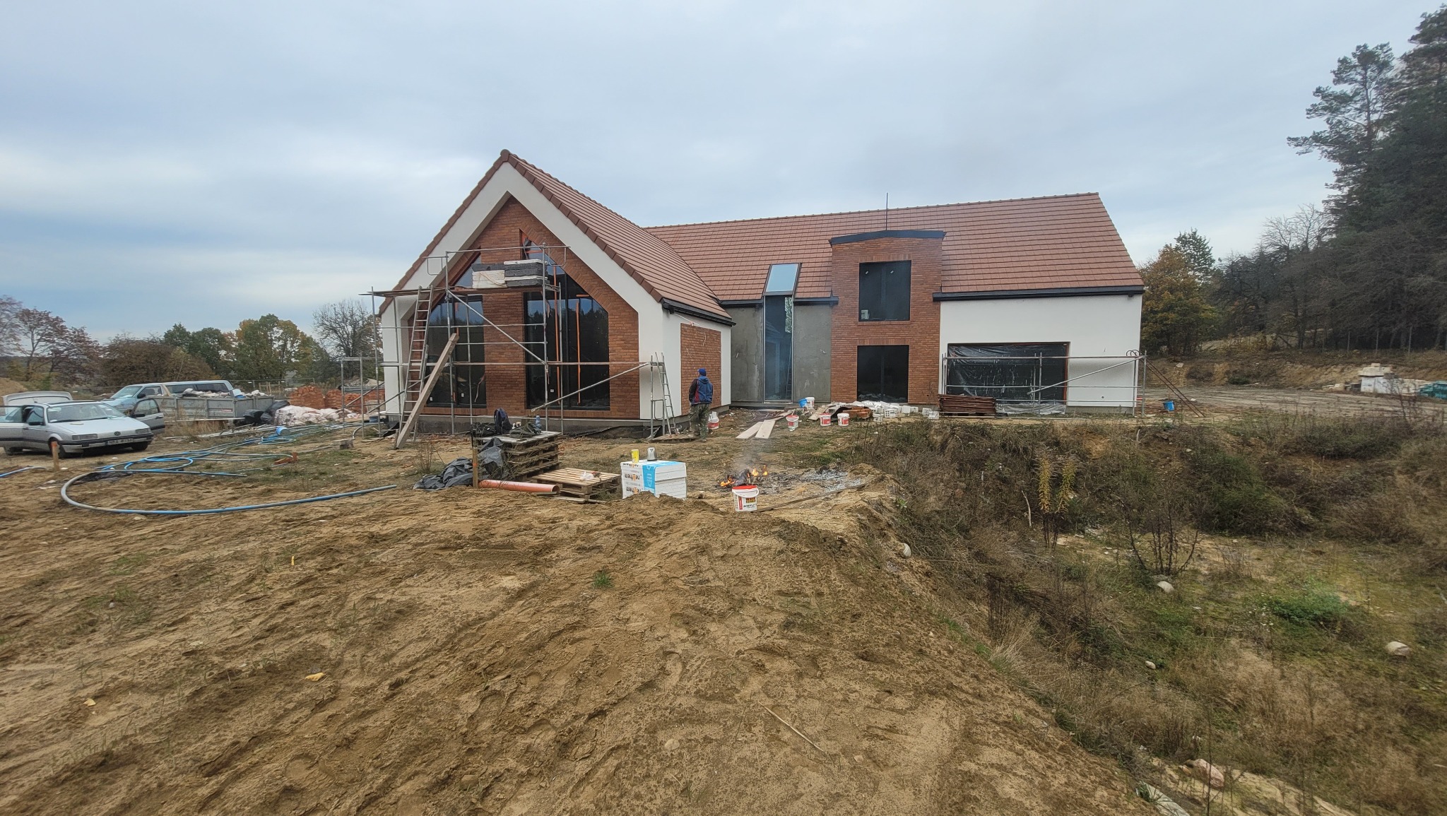 Nowoczesny dom jednorodzinny w trakcie budowy, z widoczną elewacją z cegły klinkierowej i białego tynku, częściowo osłonięty rusztowaniami, na pierwszym planie ziemia i sprzęt budowlany.