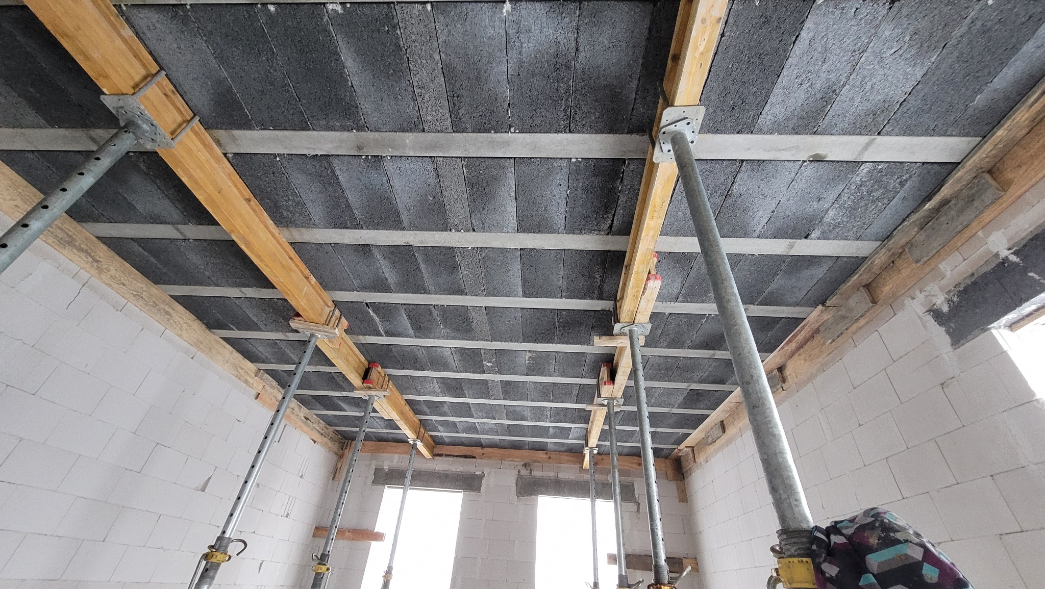 Wnętrze w trakcie budowy domu z widocznym stropem z betonowych bloczków, podpartym drewnianymi belkami i metalowymi stemplami, ściany z białych bloczków, dwa okna w tle.