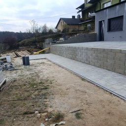 Marcin Piersa usługi budowlane - Ścieżka z szarej kostki brukowej prowadząca wzdłuż murku z cegieł, z paletą kostki w tle. Widok na dom i ogród w pochmurny dzień.