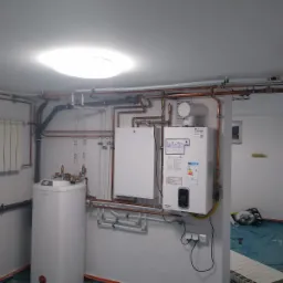 Nowa instalacja grzewcza w kotłowni: biały piec dwufunkcyjny, zasobnik ciepłej wody, miedziane rury, grzejnik panelowy, zabezpieczona podłoga folią malarską.