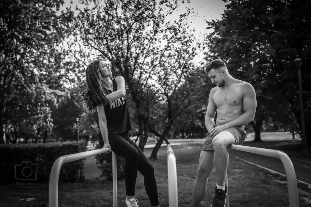 Czarno-białe zdjęcie pary siedzącej na poręczach w parku: kobieta w koszulce Nike, spoglądająca w górę, i mężczyzna bez koszulki, zamyślony, w tle drzewa.