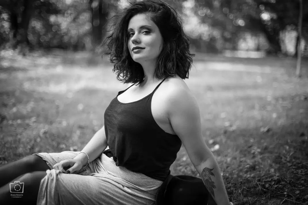 Portret kobiety siedzącej na trawie w parku, czarno-białe zdjęcie, z tatuażem łapacza snów na ramieniu i kapeluszem obok.