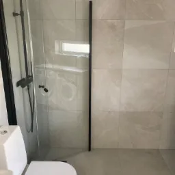 Nowoczesna łazienka z kabiną prysznicową typu walk-in z czarnymi elementami armatury, płytki w odcieniach beżu, widoczna toaleta i odbicie pralki w szklanej ściance.