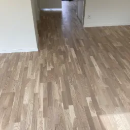 Jasne, drewniane panele ułożone w jodełkę na podłodze w nowym mieszkaniu, widoczny fragment ściany i drzwi.