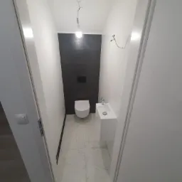 Wąska łazienka z toaletą podwieszaną, umywalką nablatową i czarną ścianą, widok zza futryny drzwiowej, surowa żarówka zamiast lampy.