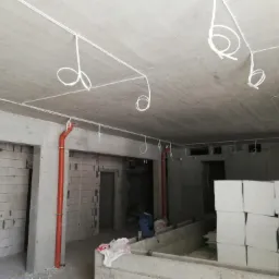 Surowy strop z widocznymi przewodami elektrycznymi w białych rurkach, zwisającymi w pętlach, oraz pionową rurą instalacyjną w kolorze pomarańczowym na tle ścian z bloczków betonowych w trakcie budowy.