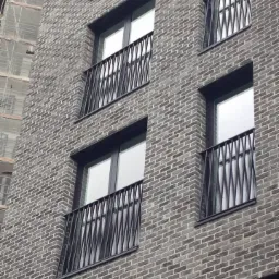 Elewacja budynku z szarej cegły z oknami zabezpieczonymi czarnymi, metalowymi balustradami o geometrycznym wzorze, widok z dołu pod kątem.