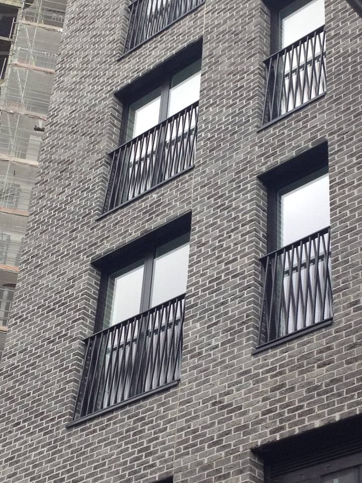 Elewacja budynku z szarej cegły z oknami zabezpieczonymi czarnymi, metalowymi balustradami o geometrycznym wzorze, widok z dołu pod kątem.