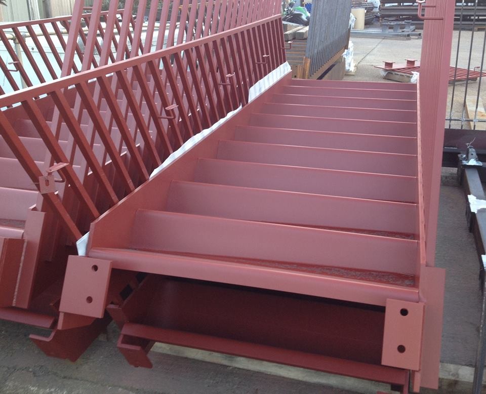 Czerwone, metalowe schody z barierką, w trakcie produkcji lub transportu, zabezpieczone białą folią, widoczne elementy mocujące.