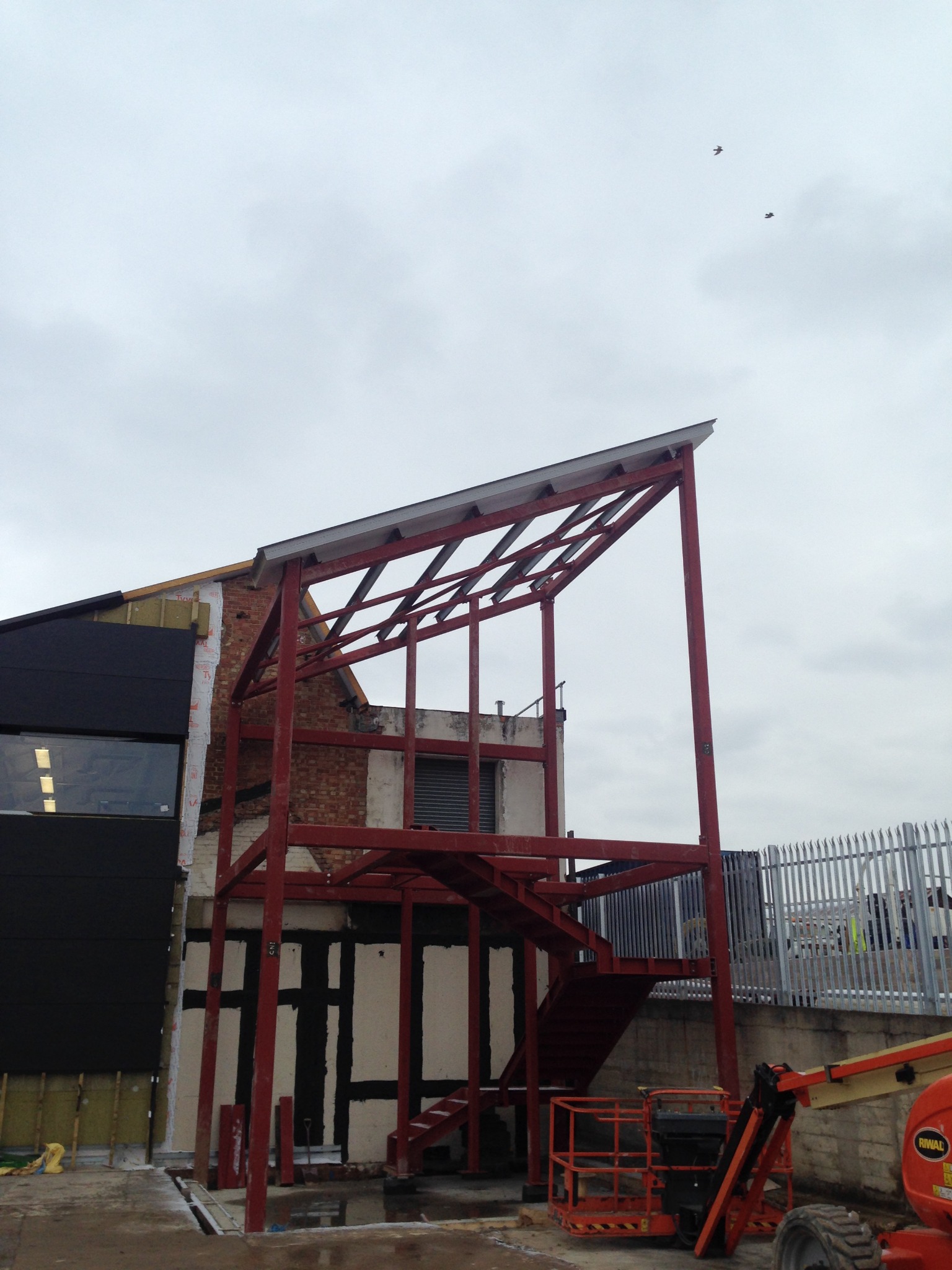 Czerwona, stalowa konstrukcja szkieletowa budynku z widocznymi schodami, częściowo obudowana, z podnośnikiem koszowym w tle i ptakami na niebie.