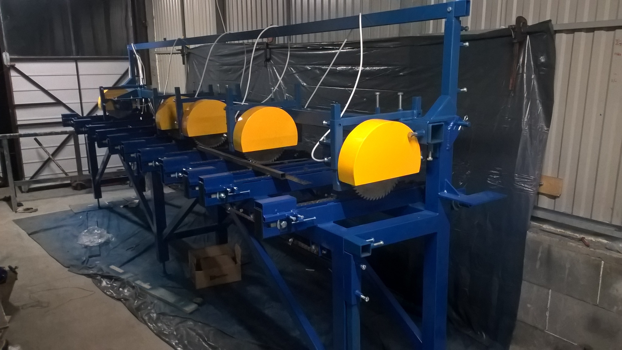 Wielopiłowa maszyna do cięcia metalu z osłonami w kolorze żółtym, rama w kolorze niebieskim, w hali produkcyjnej.