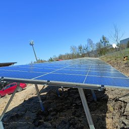 SIESTA COMFORT Szczepan Jóźwiak - Rząd paneli słonecznych na metalowej konstrukcji, z widocznym czerwonym samochodem i stertą palet w tle, na nierównym terenie z gruzem.