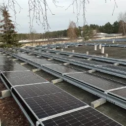 Rzędy paneli fotowoltaicznych zamontowane na płaskim dachu w Ożarowie Mazowieckim, widok z góry z fragmentem drzewa w tle.