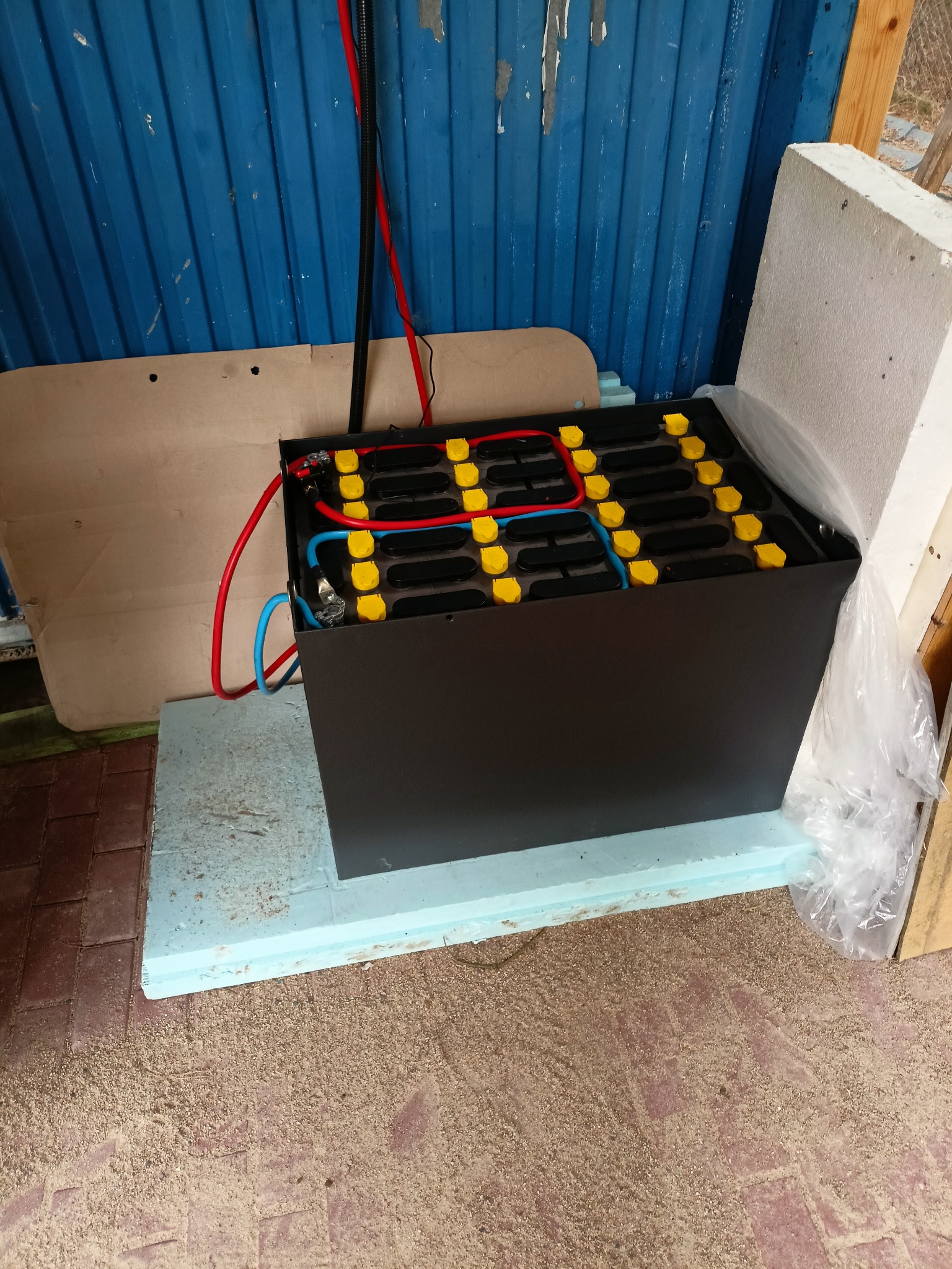 Czarna skrzynka z akumulatorami z żółtymi złączkami, połączona czerwonymi i niebieskimi kablami, stojąca na niebieskiej płycie styropianowej, na tle niebieskiej, pomalowanej blachy falistej.