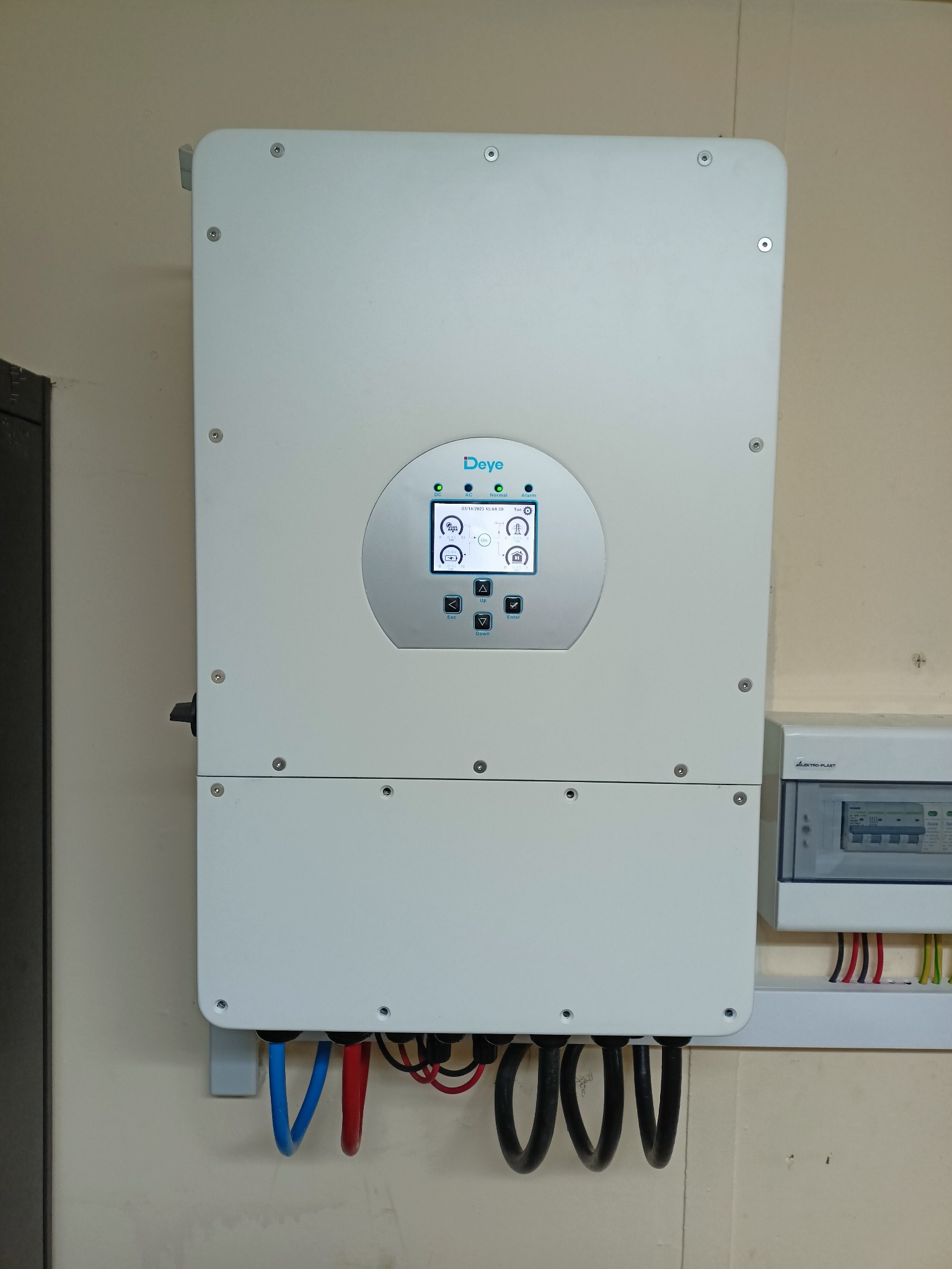 Biała skrzynka inwertera marki Deye zamontowana na ścianie, z widocznymi kablami i panelem sterowania z wyświetlaczem, obok skrzynka bezpieczników.
