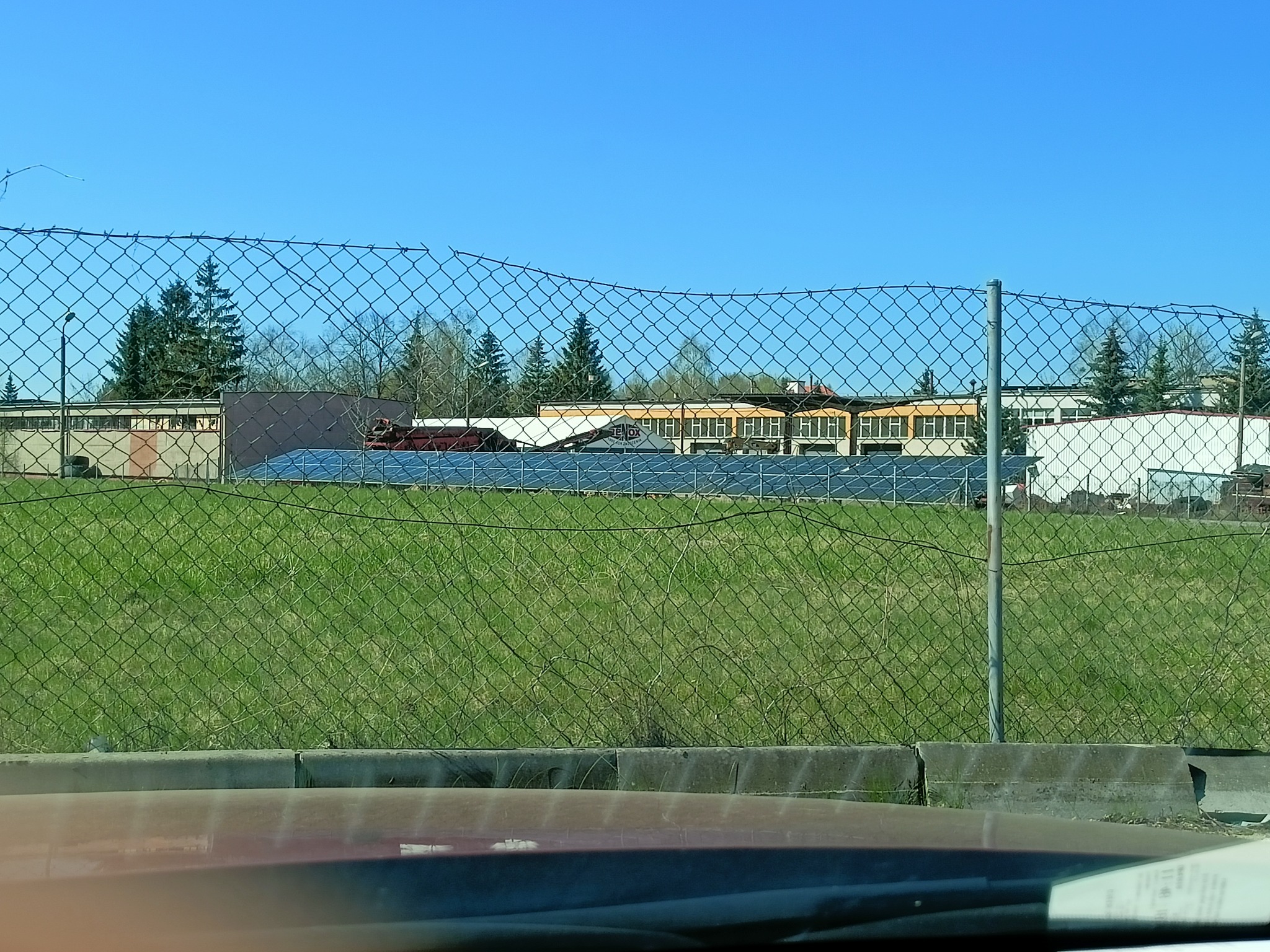 Widok instalacji fotowoltaicznej na dachu budynku przemysłowego, widziany przez ogrodzenie z siatki, w tle drzewa i błękitne niebo.