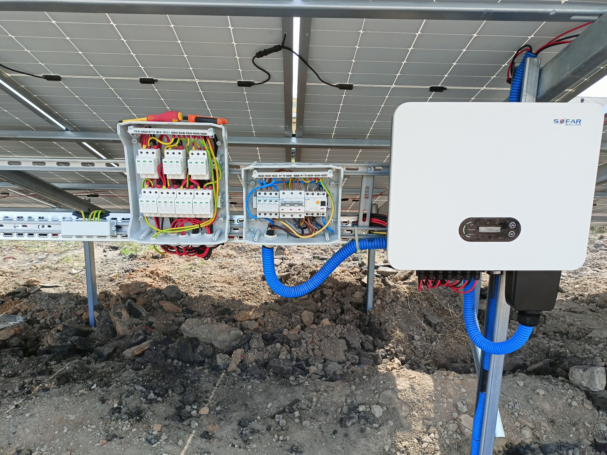 Panel fotowoltaiczny z widocznymi skrzynkami przyłączeniowymi, inwerterem Sofar Solar i niebieskimi rurami karbowanymi na grząskim terenie.