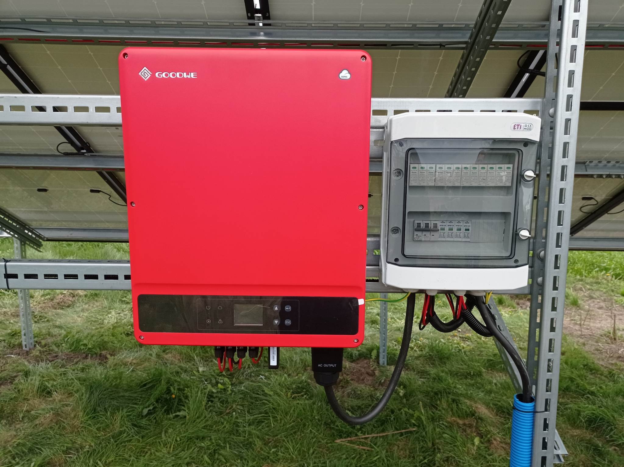 Czerwony inwerter marki GoodWe zamontowany obok skrzynki zabezpieczeń na tle paneli słonecznych i trawy.