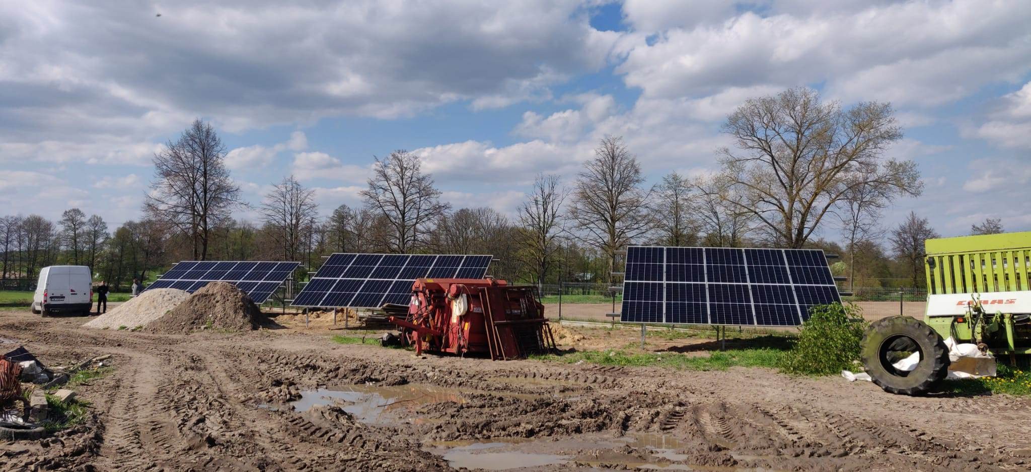 Instalacja paneli słonecznych na polu, widoczne panele, maszyny rolnicze i błotnista droga dojazdowa.
