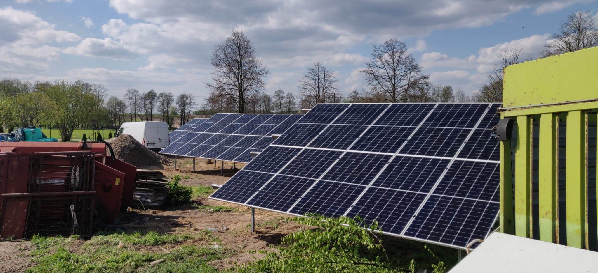 Rząd paneli słonecznych na metalowych podporach, z transportowym vanem w tle, na terenie budowy z zielonymi roślinami i sprzętem rolniczym w pierwszym planie.