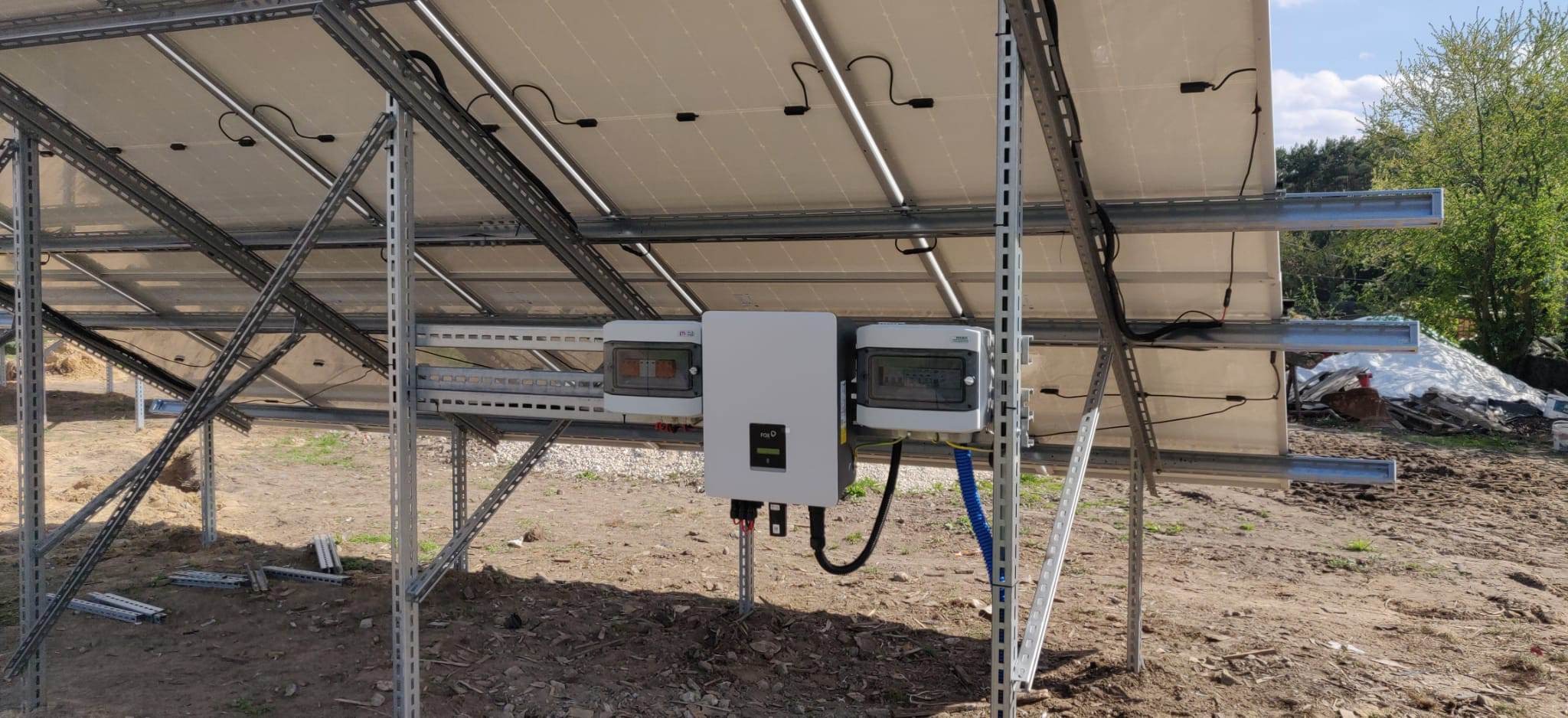 Widok od spodu na konstrukcję wsporczą paneli fotowoltaicznych z zamontowanym inwerterem i skrzynką zabezpieczeń na tle gruntu i zieleni.