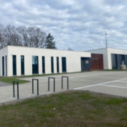 ZAKŁAD REMONTOWO - BUDOWLANY ZBIGNIEW KUBACZYK - Nowoczesny, parterowy budynek z białymi ścianami i ciemnymi, pionowymi oknami. Widoczne elementy zagospodarowania terenu i fragment parkingu. Pochmurne niebo w tle.