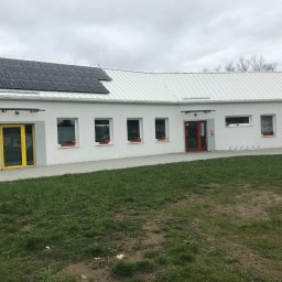 ZAKŁAD REMONTOWO - BUDOWLANY ZBIGNIEW KUBACZYK - Biały, parterowy budynek z panelami słonecznymi na dachu, kolorowymi drzwiami (żółte, czerwone) i kwiatami w oknach. Widok z zewnątrz, trawnik przed budynkiem.