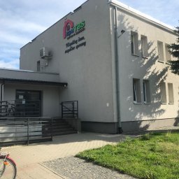ZAKŁAD REMONTOWO - BUDOWLANY ZBIGNIEW KUBACZYK - Szary budynek ZGM TBS w Szczecinku z rampą dla niepełnosprawnych, rowerem i zielenią. Widoczne logo firmy i polska flaga. Słoneczny dzień.