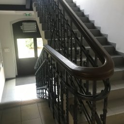 ZAKŁAD REMONTOWO - BUDOWLANY ZBIGNIEW KUBACZYK - Klatka schodowa z metalową, ozdobną balustradą i szarymi schodami. Widoczne płytki na podłodze i drzwi z napisem 'exit'.