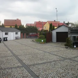 Obszerny podjazd wyłożony kostką granitową w geometryczny wzór, z widocznymi budynkami mieszkalnymi w tle.