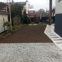 DAWID WIĘZIK - Przygotowany teren pod trawnik z nowo ułożonym chodnikiem z jasnych kamieni i płyt betonowych, widoczna zagęszczarka w tle, huśtawka ogrodowa.