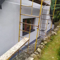 Świeżo pomalowana elewacja domu w odcieniach szarości, widoczna z bliska, z żółtym rusztowaniem przylegającym do ściany i kamiennym obrzeżem wzdłuż trawnika.