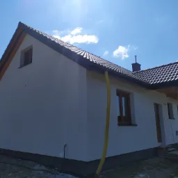 Jasnoszary dom jednorodzinny z ciemną dachówką, widok z boku, żółty wąż wentylacyjny przy elewacji, błękitne niebo z chmurami w tle.