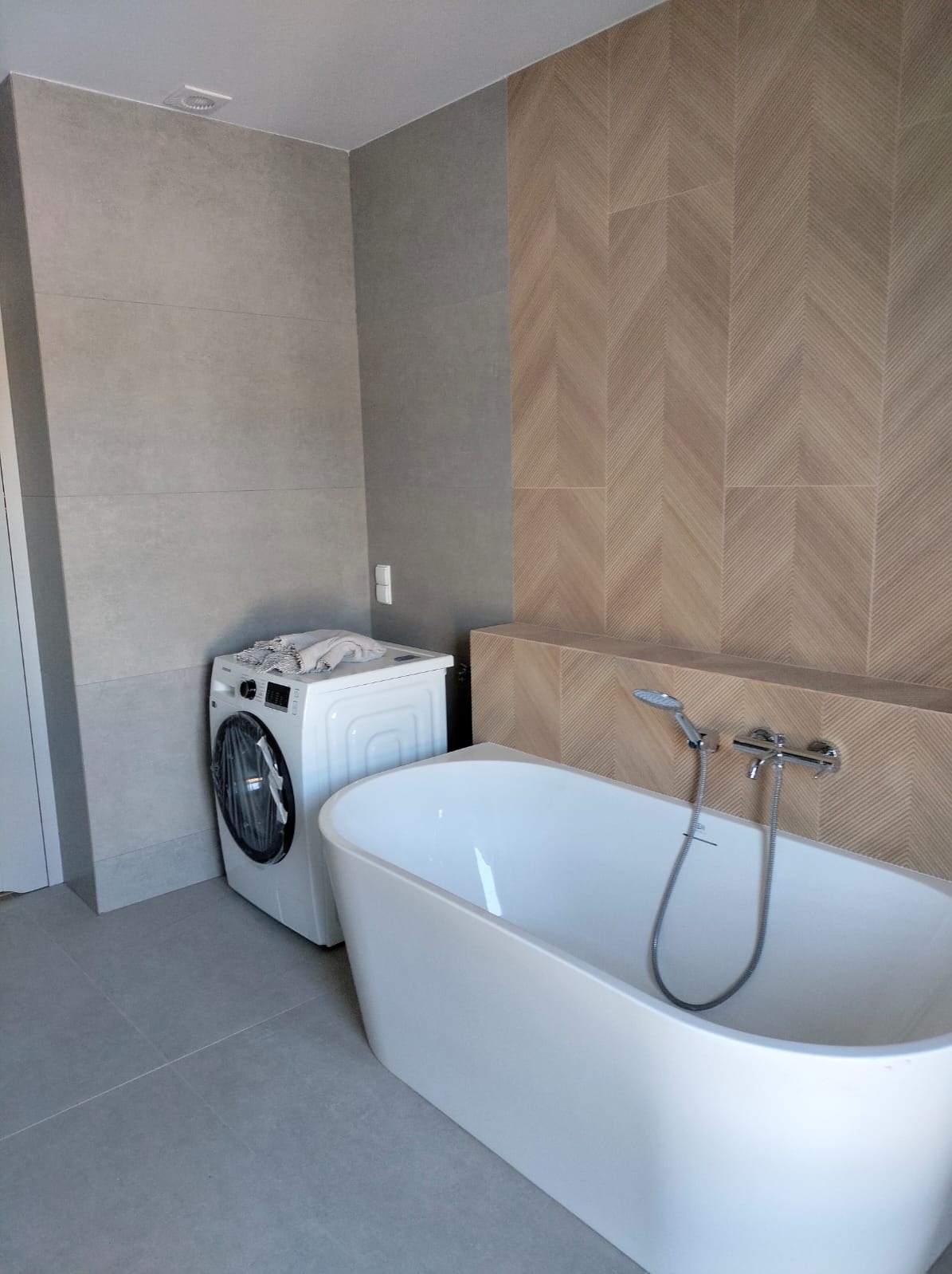 Nowoczesna łazienka z wolnostojącą białą wanną, chromowaną armaturą i pralką w rogu. Na ścianie szare płytki oraz dekoracyjne płytki ułożone we wzór jodełki.