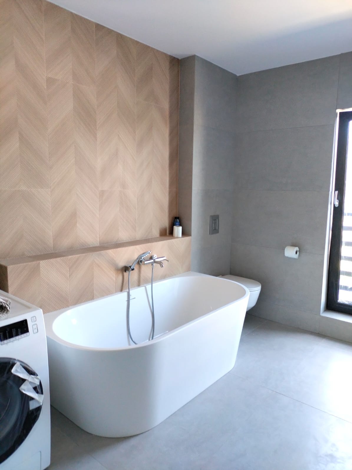 Nowoczesna łazienka z białą wanną wolnostojącą, baterią wannową, pralką i ścianą wyłożoną płytkami w jodełkę, szara podłoga i ściany.