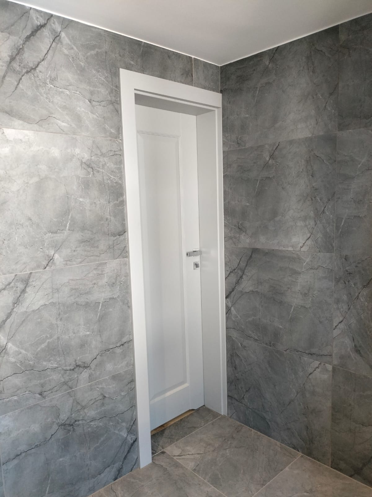 Wnętrze łazienki z szarymi płytkami imitującymi marmur na ścianach i podłodze, białe drzwi z ościeżnicą i srebrną klamką, nowoczesny design.