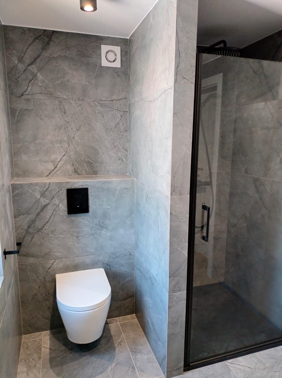 Nowoczesna łazienka z szarymi płytkami imitującymi marmur, wisząca toaleta z czarnym przyciskiem spłukującym, kabina prysznicowa z czarną ramą i deszczownicą, oraz wentylator w suficie.