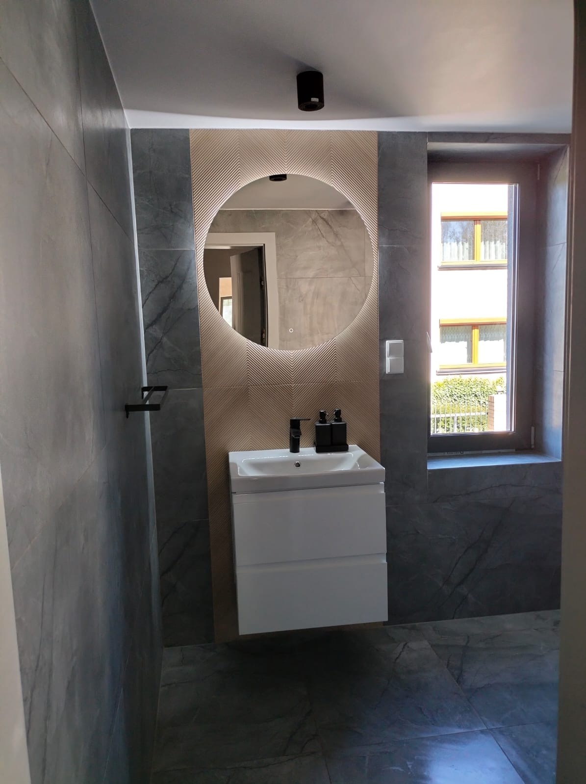 Nowoczesna łazienka w Szczyrku z szarymi płytkami imitującymi marmur, okrągłym lustrem z podświetleniem LED, białą szafką pod umywalką i czarnymi dozownikami na mydło.