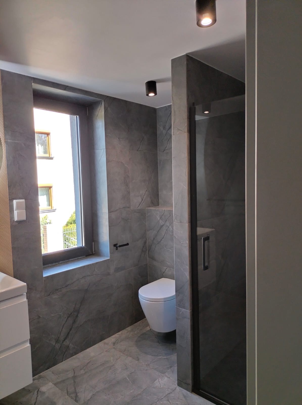 Nowoczesna łazienka z szarymi płytkami imitującymi marmur, podwieszana toaleta, kabina prysznicowa z czarnymi profilami, okno z widokiem na zewnątrz i punktowe oświetlenie sufitowe.
