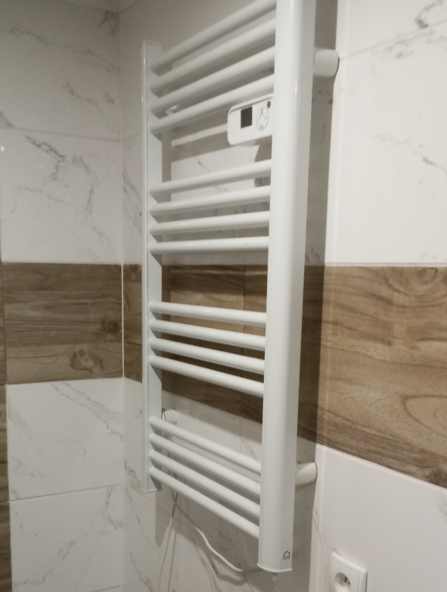 Biały, elektryczny grzejnik łazienkowy drabinkowy z termostatem zamontowany na ścianie z płytek imitujących marmur i drewno.