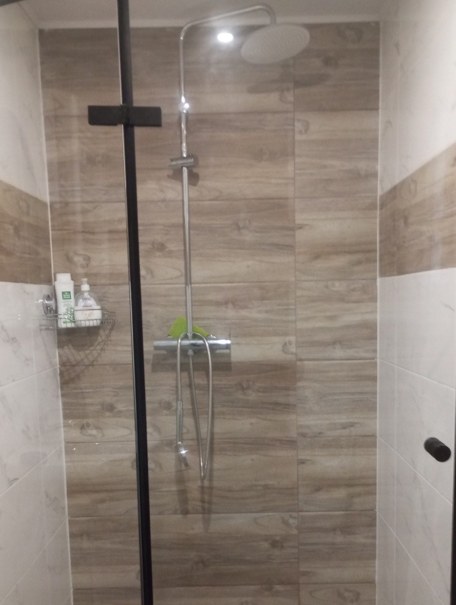 Nowoczesna kabina prysznicowa z deszczownicą i panelami ściennymi imitującymi drewno, wyposażona w półkę na kosmetyki i zieloną gąbkę.