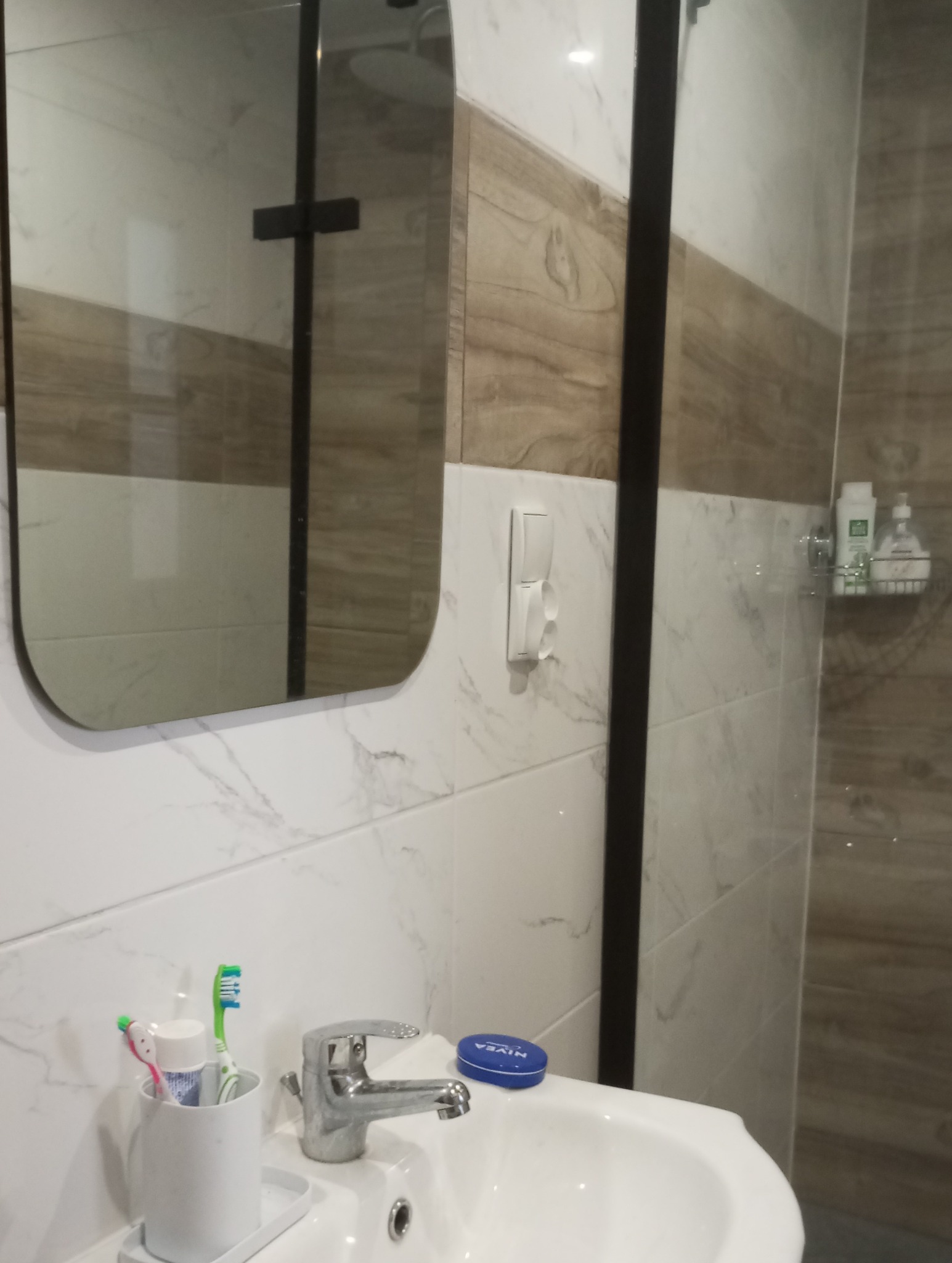 Nowoczesna łazienka z umywalką, baterią, lustrem z czarną ramą i marmurowymi płytkami w odcieniach bieli i brązu. Widoczne akcesoria łazienkowe: szczoteczki do zębów, kosmetyki i dozownik mydła.