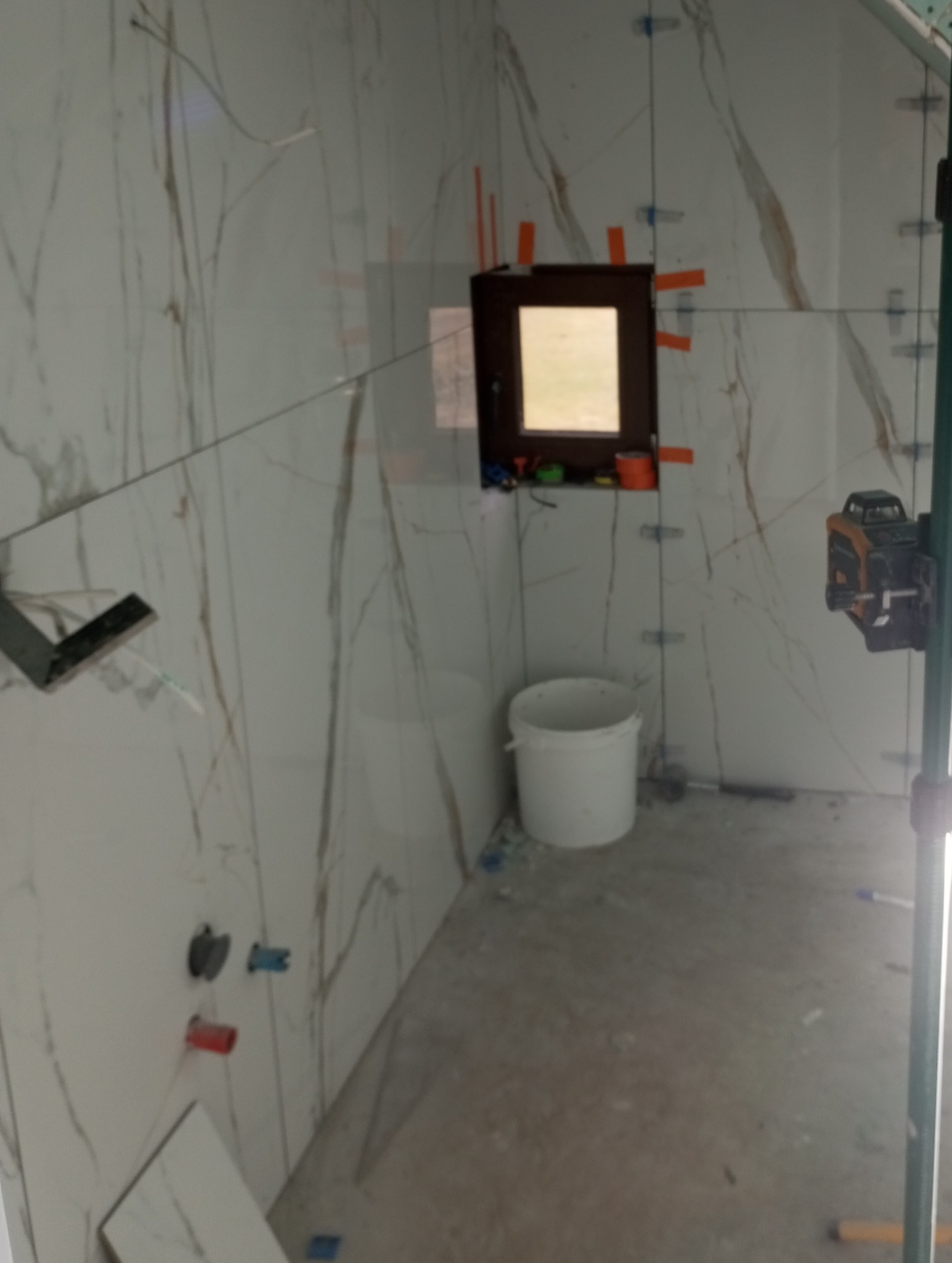 Wnętrze łazienki w trakcie wykańczania, widoczne płytki imitujące marmur z brązowymi żyłami, poziomica laserowa, wiadro i akcesoria do układania płytek.