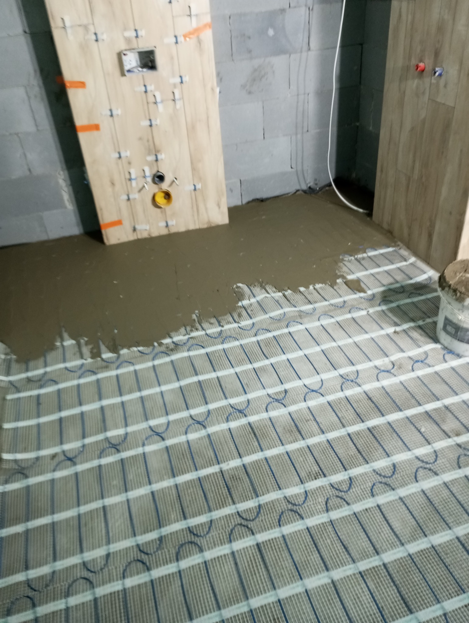 Montaż ogrzewania podłogowego elektrycznego w łazience, częściowo zalany wylewką samopoziomującą, widoczne płytki imitujące drewno na ścianie
