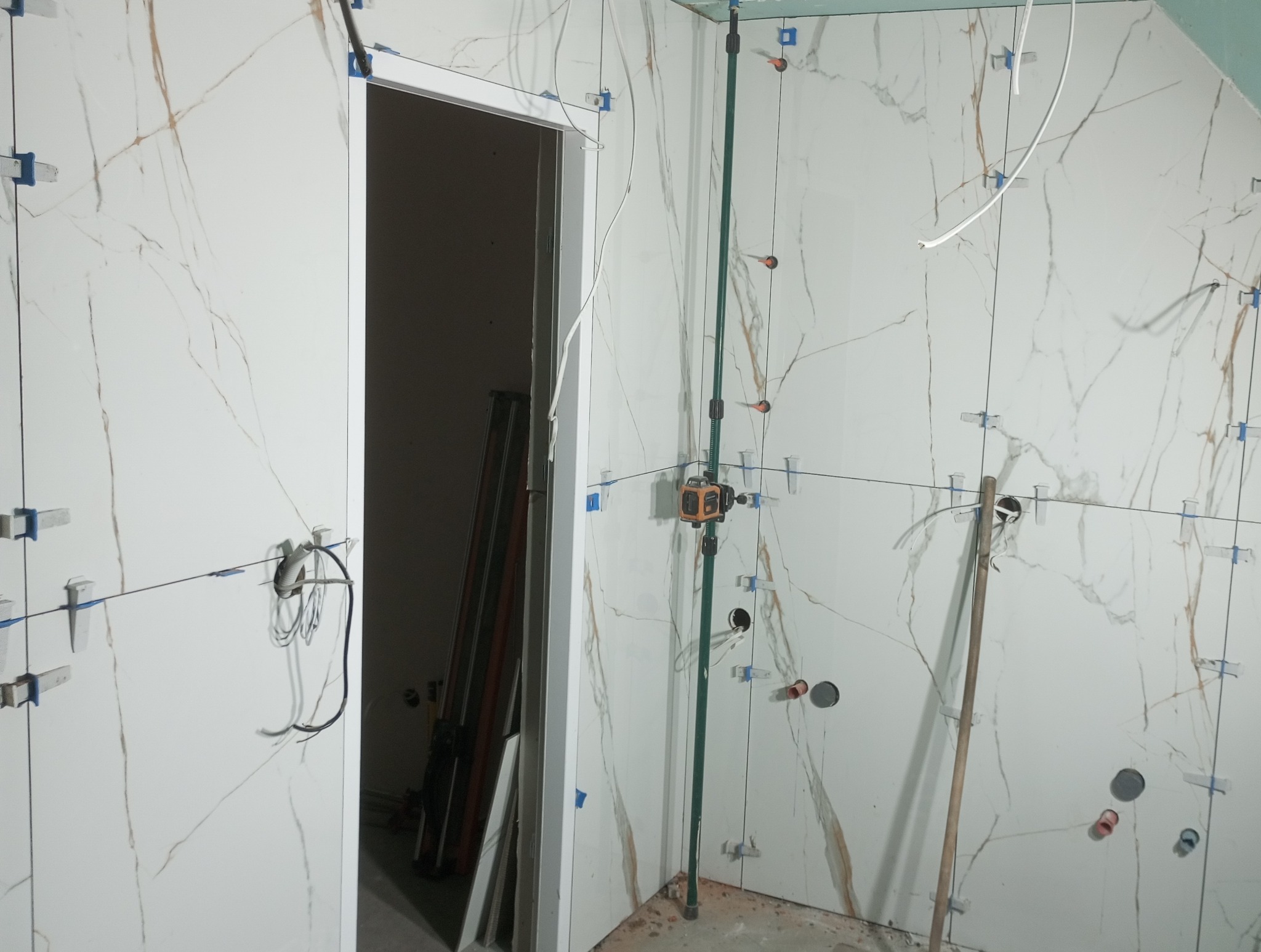 Ściana w trakcie wykańczania płytkami imitującymi marmur, z widocznymi narzędziami do układania, poziomica laserowa na statywie, otwory na gniazdka elektryczne i kable, listwy wykończeniowe przy...