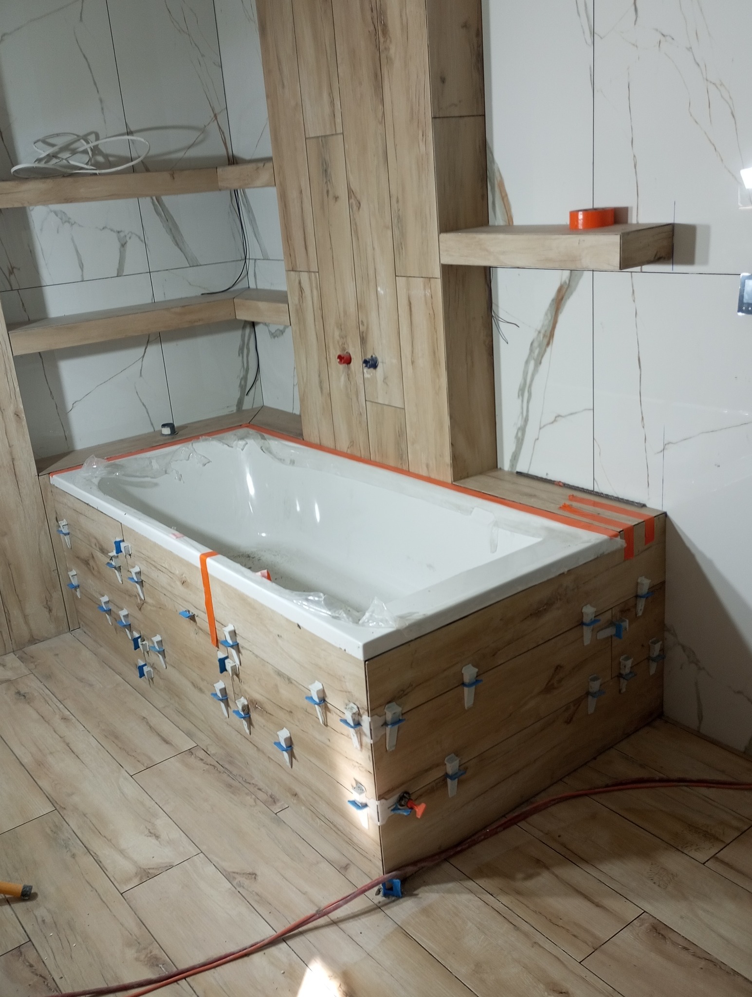 Montaż płytek imitujących drewno na obudowie wanny, z użyciem klipsów poziomujących; w tle półki z płytkami imitującymi marmur.