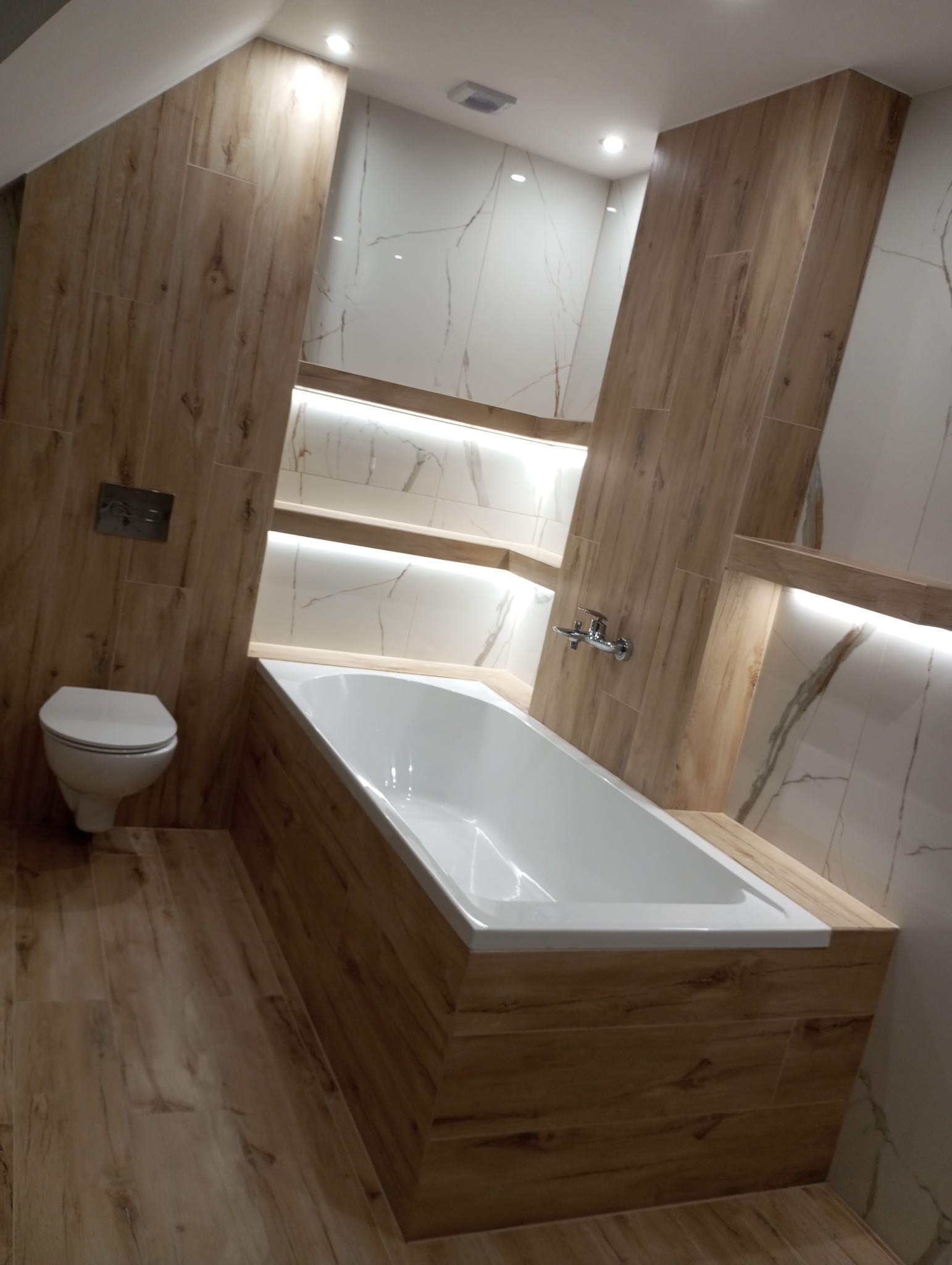 Nowoczesna łazienka z wanną obudowaną płytkami imitującymi drewno, podświetlane półki wnękowe i marmurowe płytki na ścianach